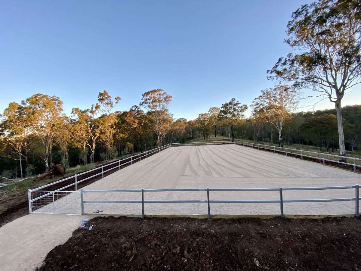 A large, fenced sand arena surrounded by trees under a clear sky at dusk. A large, fenced sand arena surrounded by trees under a clear sky at dusk.