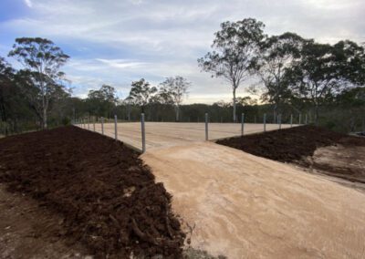 A newly constructed dirt road flanked by mounds of soil and metal poles, with trees in the background
