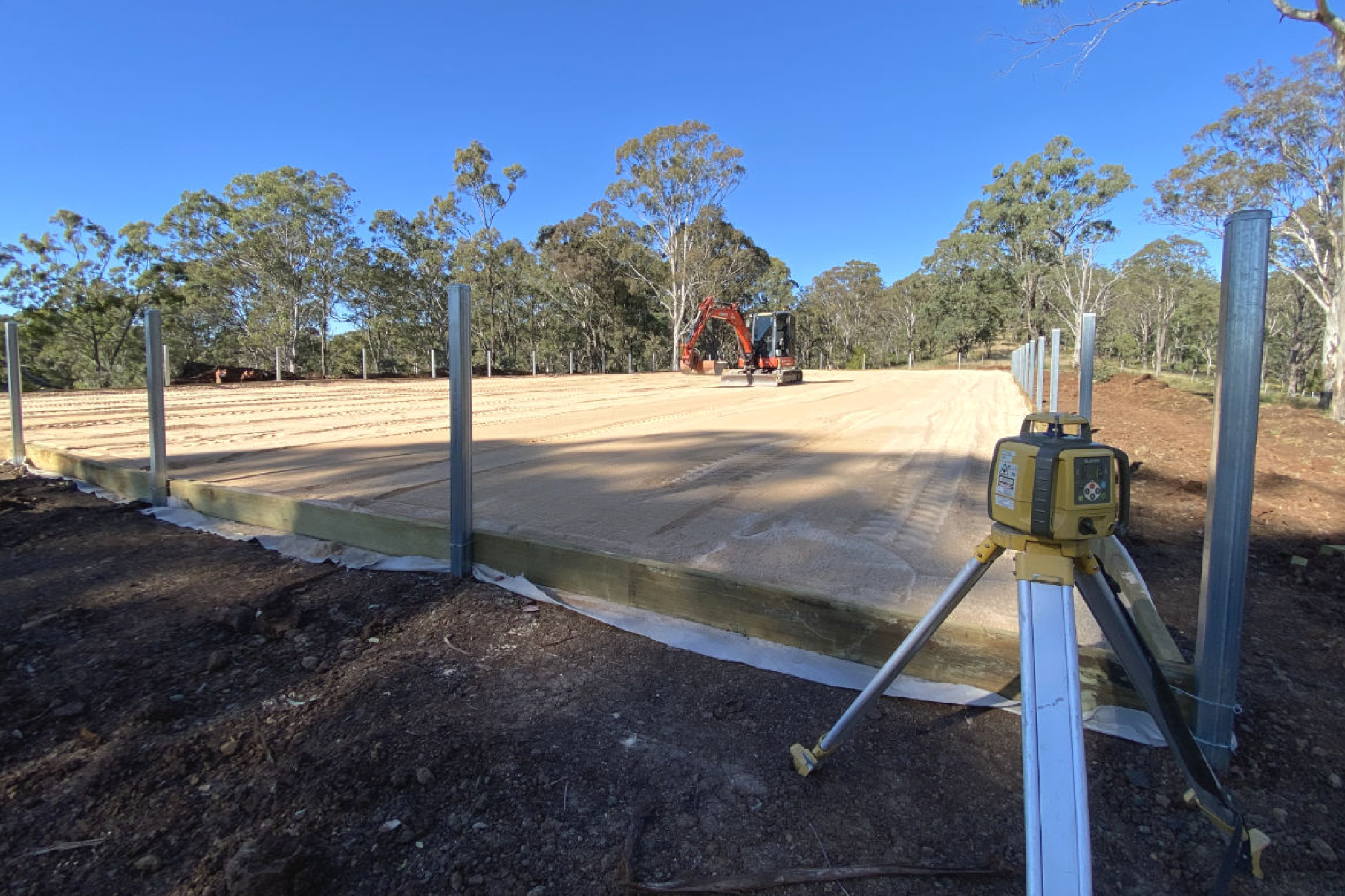 A surveyor's level in the foreground overlooking a construction site with a red excavator and new fencing