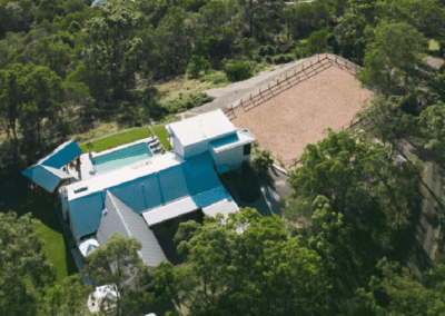 Aerial view of a modern blue-roofed building near a sandy horse arena amidst greenery