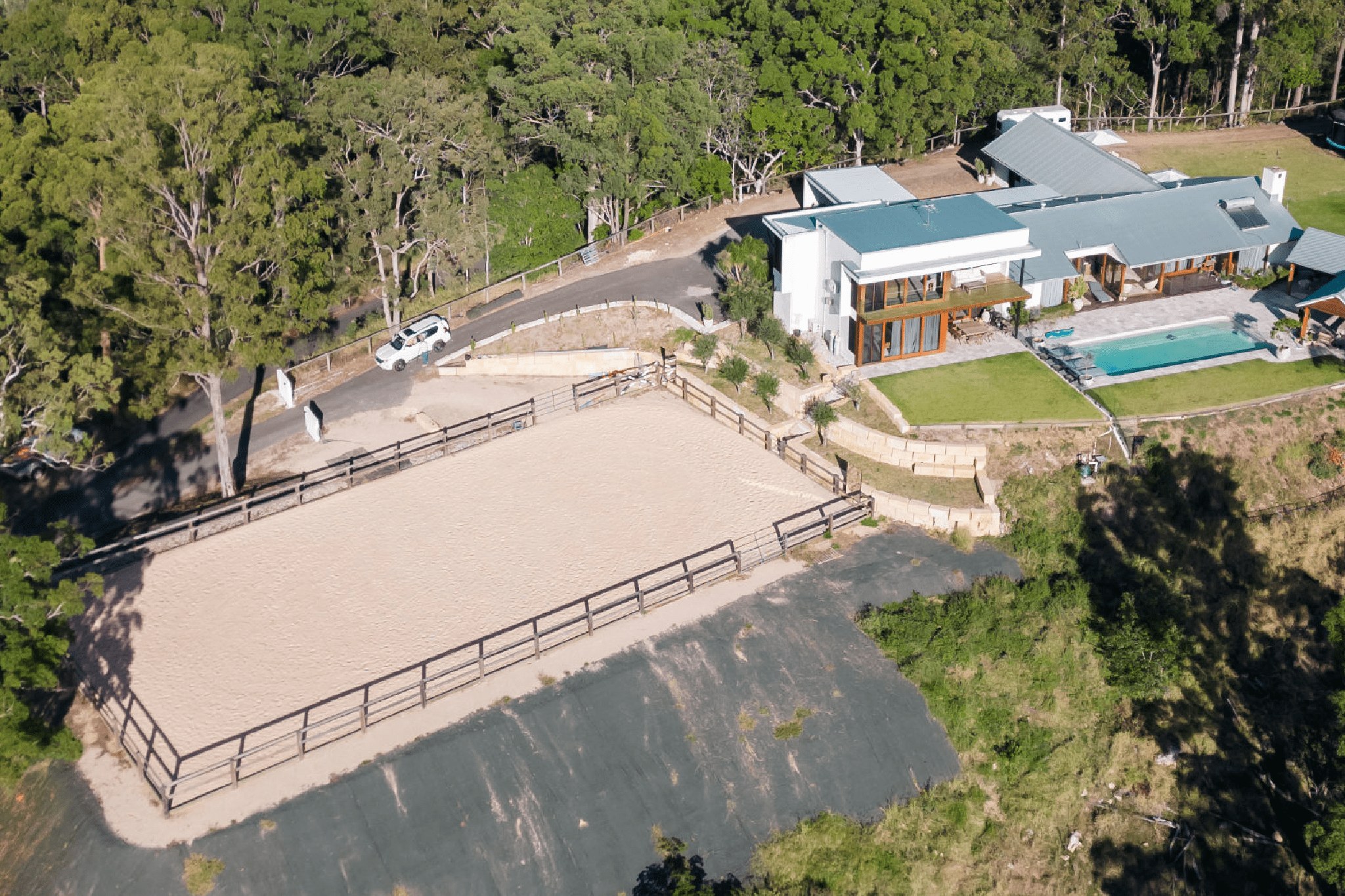 Aerial view of a modern house with a pool and adjacent horse riding arena surrounded by trees