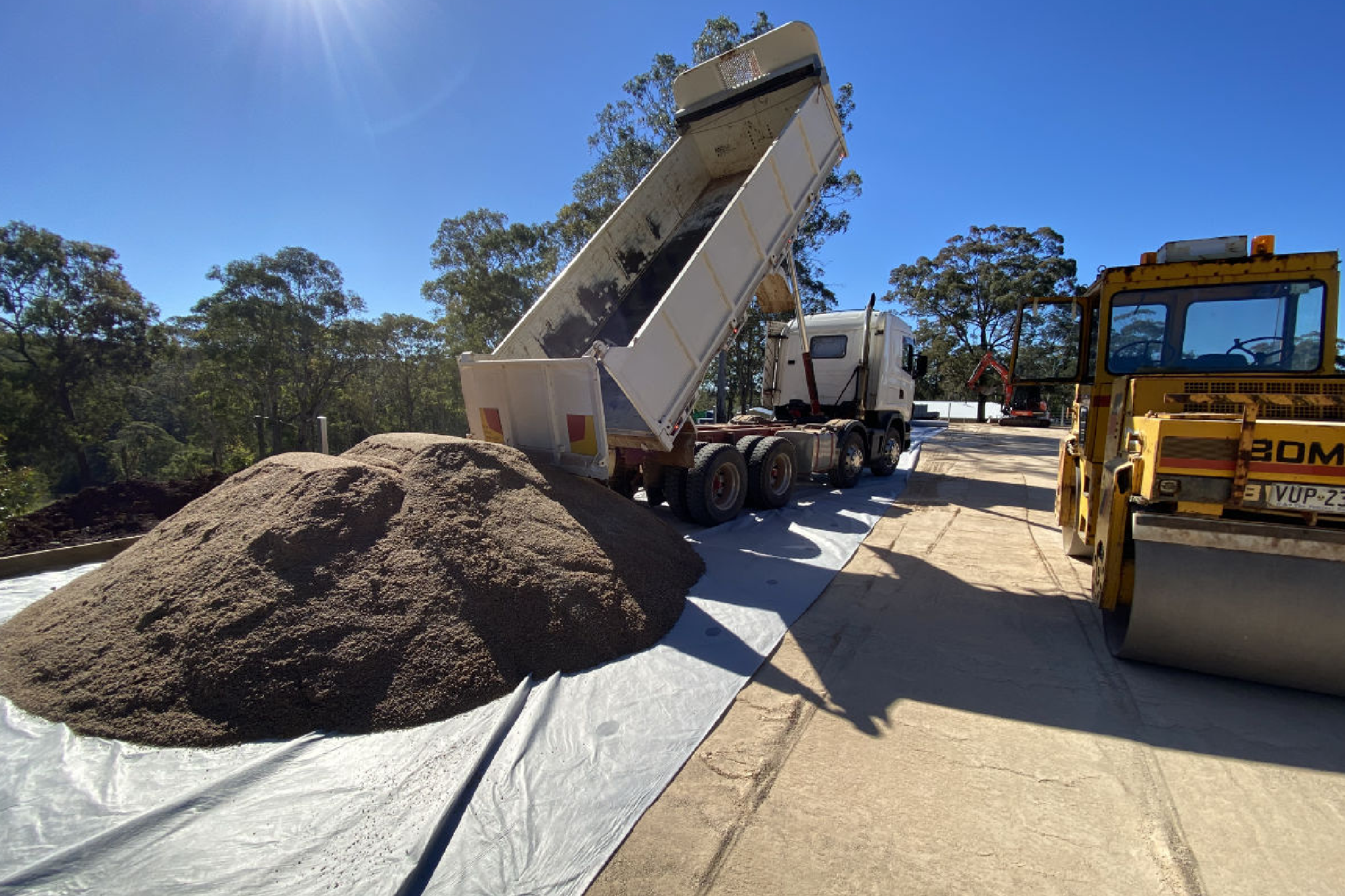 Dump truck tilting bed to pour soil next to a bulldozer, under sunny sky