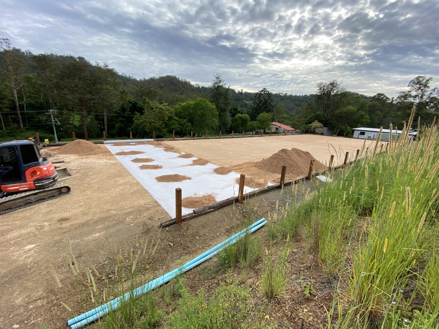 A construction site with a leveled dirt area, a red excavator, and a cloudy sky in the background.