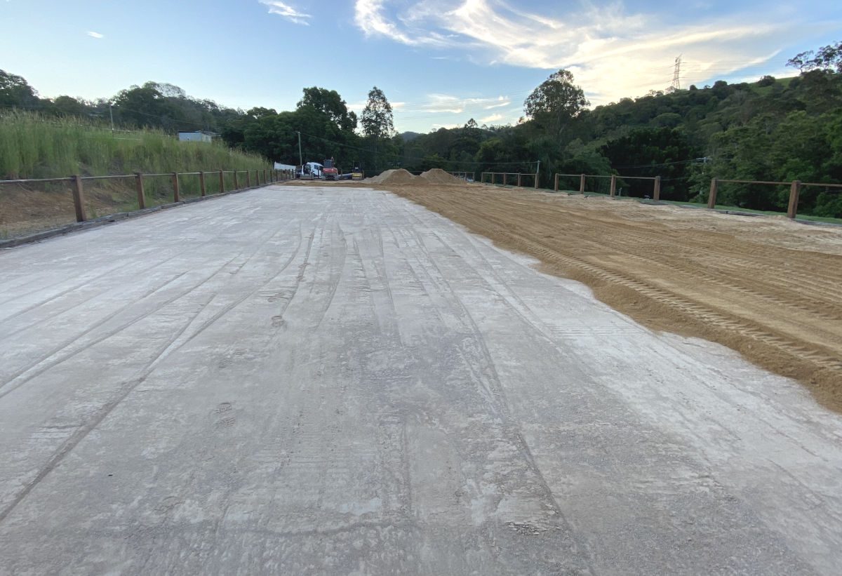 Newly constructed concrete road base with adjacent sandy area and trees under a clear sky